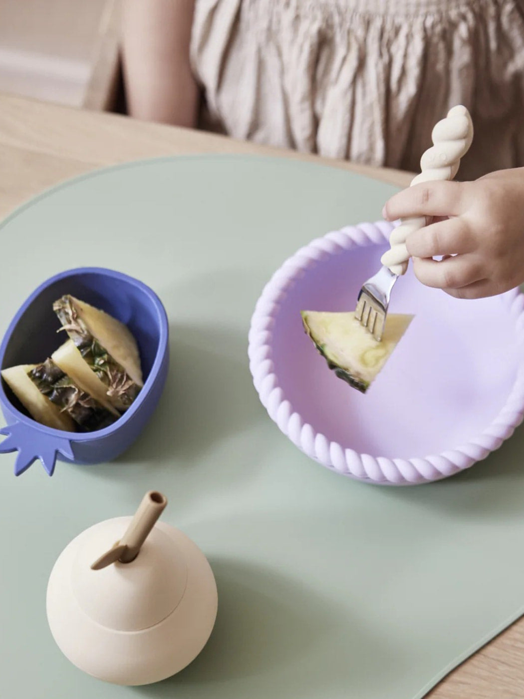 Child eating a slice of pineapple with a fork on a purple plate, next to a blue bowl with a sandwich on a light green table.