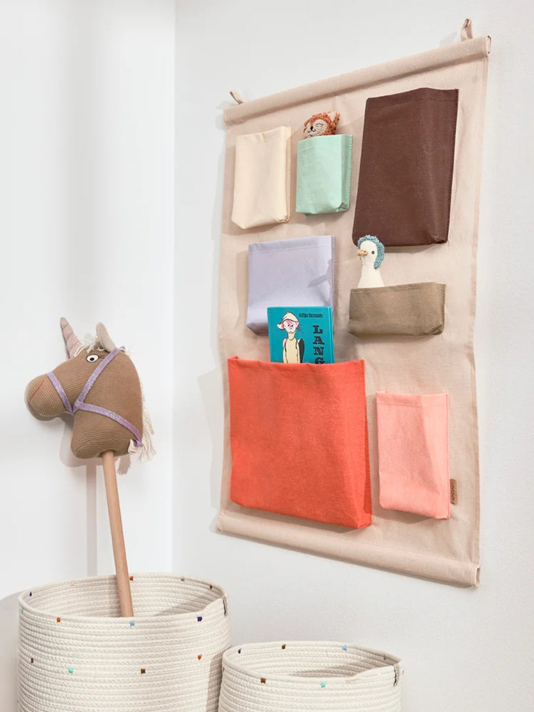 Wall-mounted fabric organizer with colorful pockets holding toys and books, next to woven baskets on a white wall.